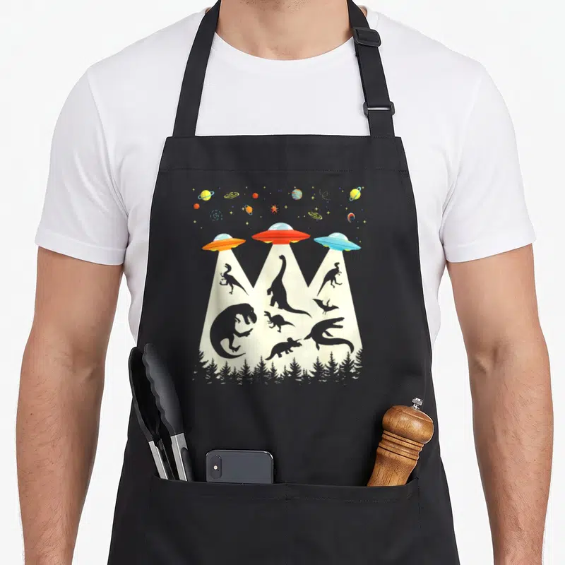 Tablier de cuisine noir avec illustration retro de dinosaures emportés par des rayons de vaisseaux spatiaux colorés au-dessus d'une forêt.
