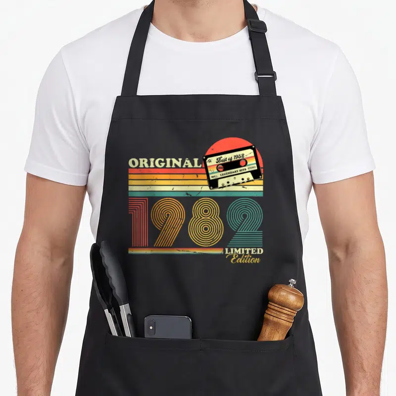Tablier de cuisine noir avec une illustration de cassette 1982 et des bandes colorées rétro sur fond uni.