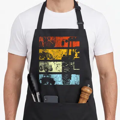 Tablier de cuisine noir avec quatre bandes horizontales colorées affichant des silhouettes de tracteurs rétro et des détails de pneus texturés.