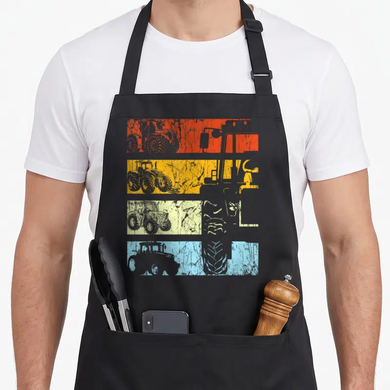 Tablier de cuisine noir avec quatre bandes horizontales colorées affichant des silhouettes de tracteurs rétro et des détails de pneus texturés.