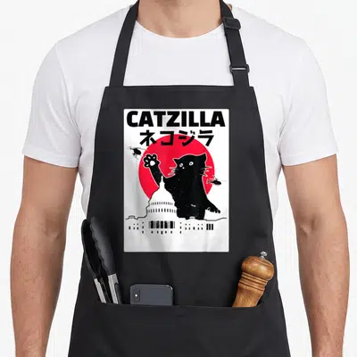 Tablier de cuisine noir avec illustration d'un chat géant noir sur fond de soleil rouge et silhouettes urbaines blanches.