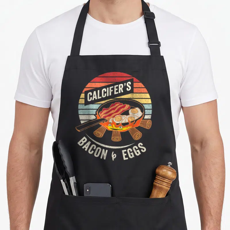 Tablier de cuisine noir avec illustration rétro d'une poêle contenant du bacon et des œufs sur fond de soleil coloré.