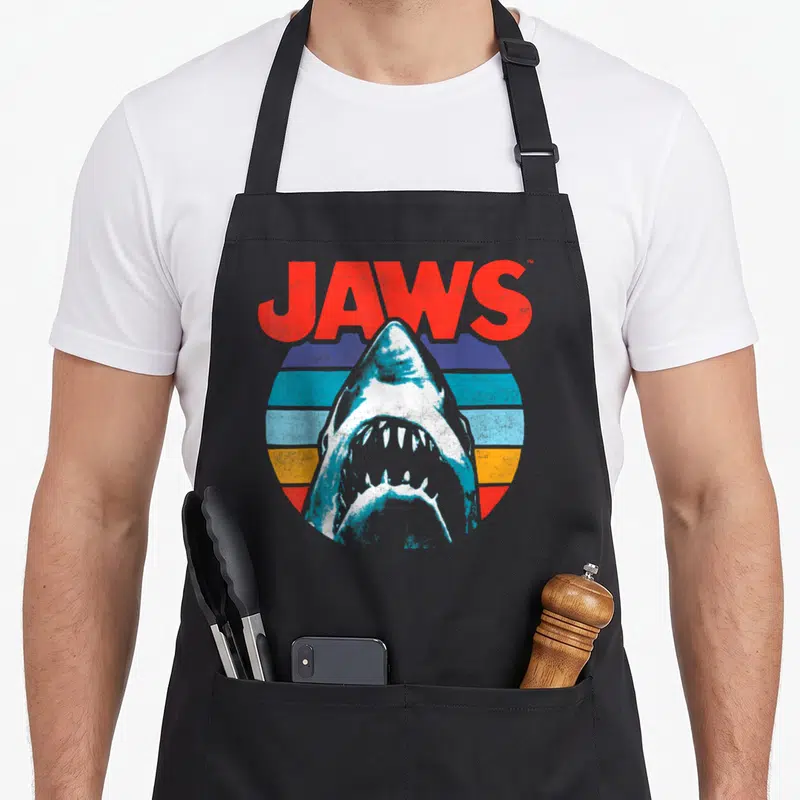 Tablier de cuisine noir avec illustration d'un requin blanc sur fond de cercle coloré et texte Jaws en rouge.