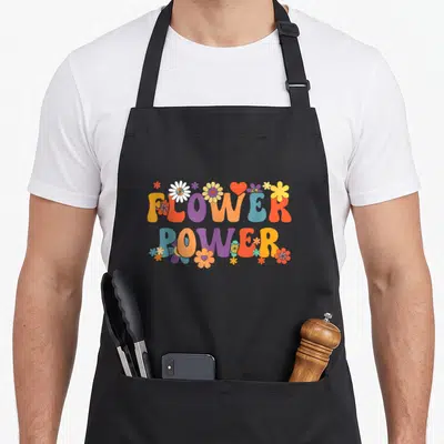 Tablier de cuisine noir avec l'inscription Flower Power entourée de fleurs multicolores et d'un cœur rouge dans un style rétro.