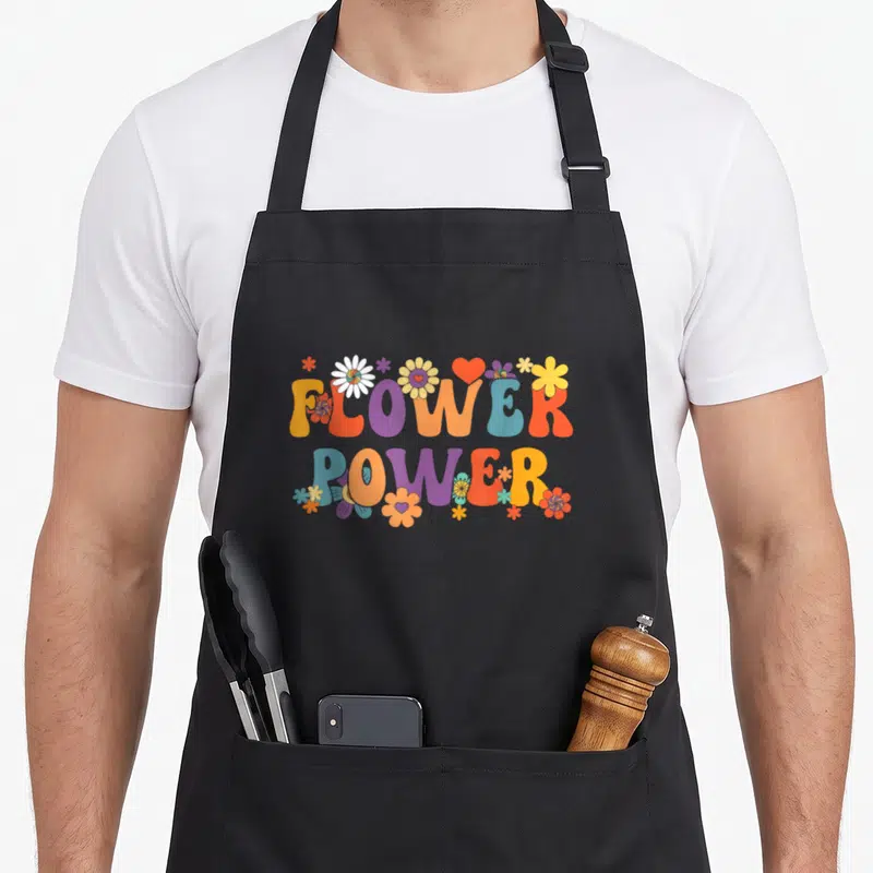 Tablier de cuisine noir avec l'inscription Flower Power entourée de fleurs multicolores et d'un cœur rouge dans un style rétro.