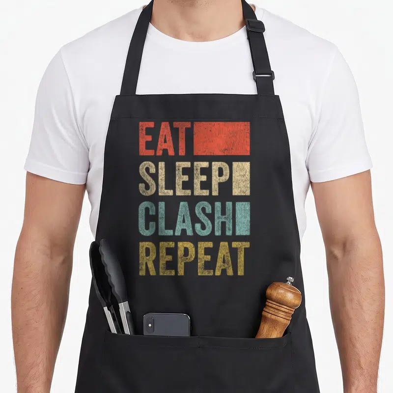 Tablier de cuisine noir avec le texte Eat Sleep Clash Repeat Clans en lettres colorées et effet vieilli.