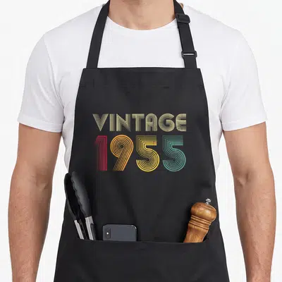 Tablier de cuisine noir avec inscription VINTAGE 1955 en lettres rayées multicolores style années cinquante.