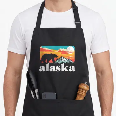 Tablier de cuisine noir avec illustration rétro d'un ours face aux montagnes d'Alaska et un ciel coloré.