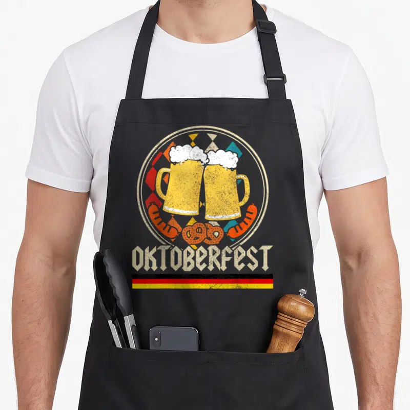 Tablier de cuisine noir avec deux gobelets de bière en toast, saucisses et texte Oktoberfest rétro sur fond circulaire coloré.