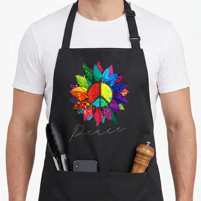 Tablier de cuisine noir avec un grand tournesol multicolore et un signe de la paix au centre, signé Peace en bas.