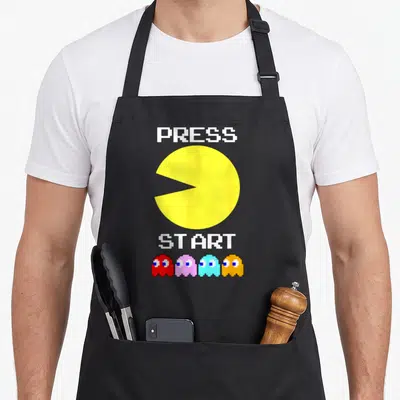 Tablier de cuisine noir avec un grand Pac-Man jaune, des fantômes colorés et le texte PRESS START en blanc pixelisé.