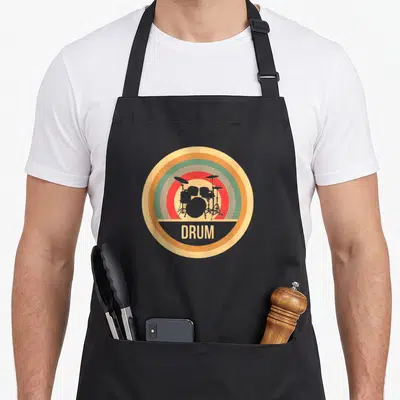 Tablier de cuisine noir avec un logo circulaire rétro illustrant une batterie et le mot DRUM en lettres capitales beige.