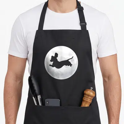 Tablier de cuisine noir avec silhouette de chien teckel sautant devant une lune texturée en nuances de gris.