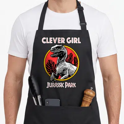 Tablier de cuisine noir avec illustration d'un raptor rouge et blanc encadré par le texte Clever Girl Jurassic Park.
