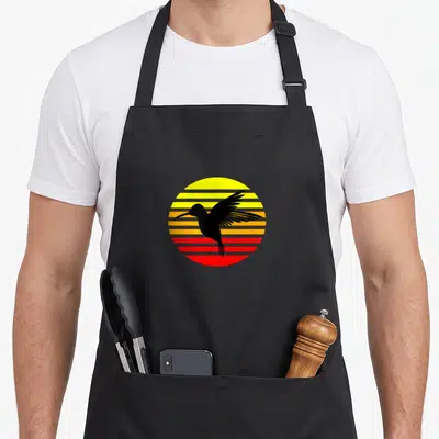 Tablier de cuisine noir avec illustration d'un colibri volant devant un soleil rayé jaune orange et rouge au design rétro.