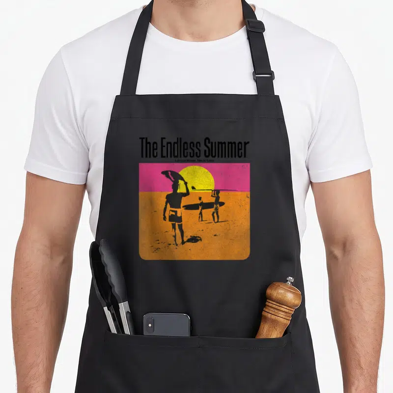 Tablier de cuisine noir avec illustration retroThe Endless Summer montrant des surfeurs et un soleil jaune sur fond rose et orange.