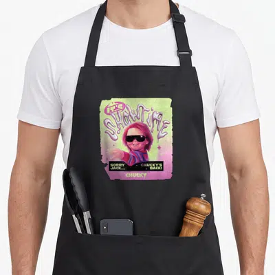 Tablier de cuisine noir avec une illustration rétro de la poupée Chucky aux cheveux roses et lunettes noires.
