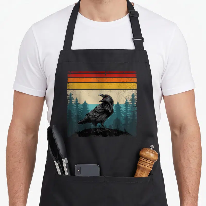 Tablier de cuisine noir avec illustration corbeau rétro sur fond de pins et bandes colorées orange jaune rouge