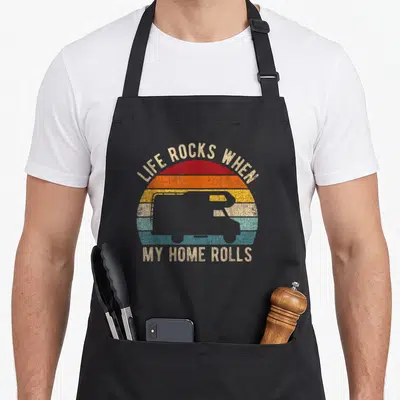 Tablier de cuisine noir avec illustration camping-car rétro et texte Life Rocks When My Home Rolls sur fond de soleil coloré.