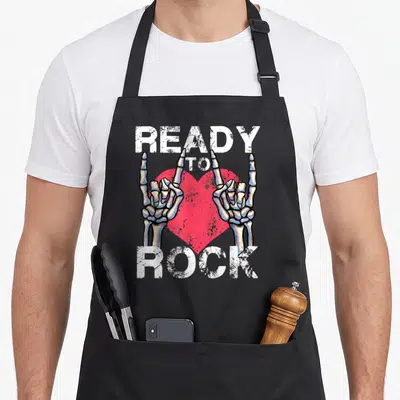 Tablier de cuisine noir avec deux mains squelettiques formant le signe du rock devant un cœur rose et du texte blanc délavé.
