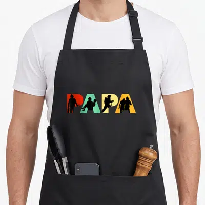 Tablier de cuisine noir avec linscription PAPA en lettres multicolores intégrant des silhouettes de pompiers en action.