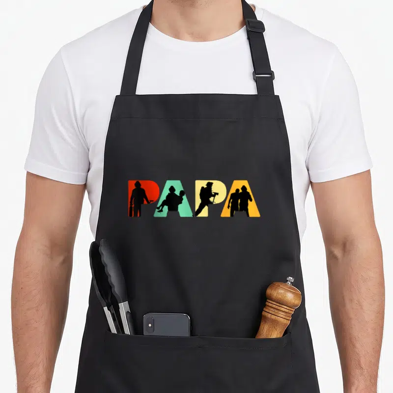 Tablier de cuisine noir avec l'inscription PAPA en lettres multicolores intégrant des silhouettes de pompiers en action.