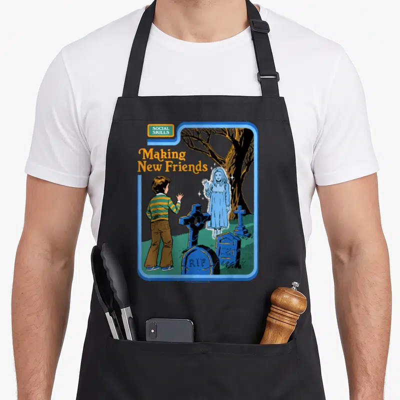 Tablier de cuisine noir avec illustration rétro d'un enfant saluant un fantôme bleu dans un cimetière sous l'inscription Making New Friends.