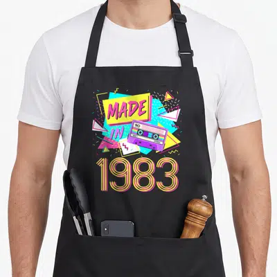 Tablier de cuisine noir avec illustration d'une cassette rose et le texte 1983 en lettres rayées jaunes et roses.