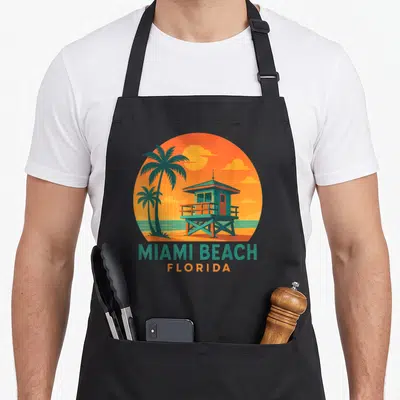 Tablier de cuisine noir avec illustration circulaire orange et turquoise représentant une cabane de sauveteur et des palmiers à Miami Beach Florida.