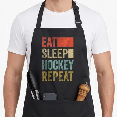 Tablier de cuisine noir avec le texte Eat Sleep Hockey Repeat en lettres colorées et effet vieilli.