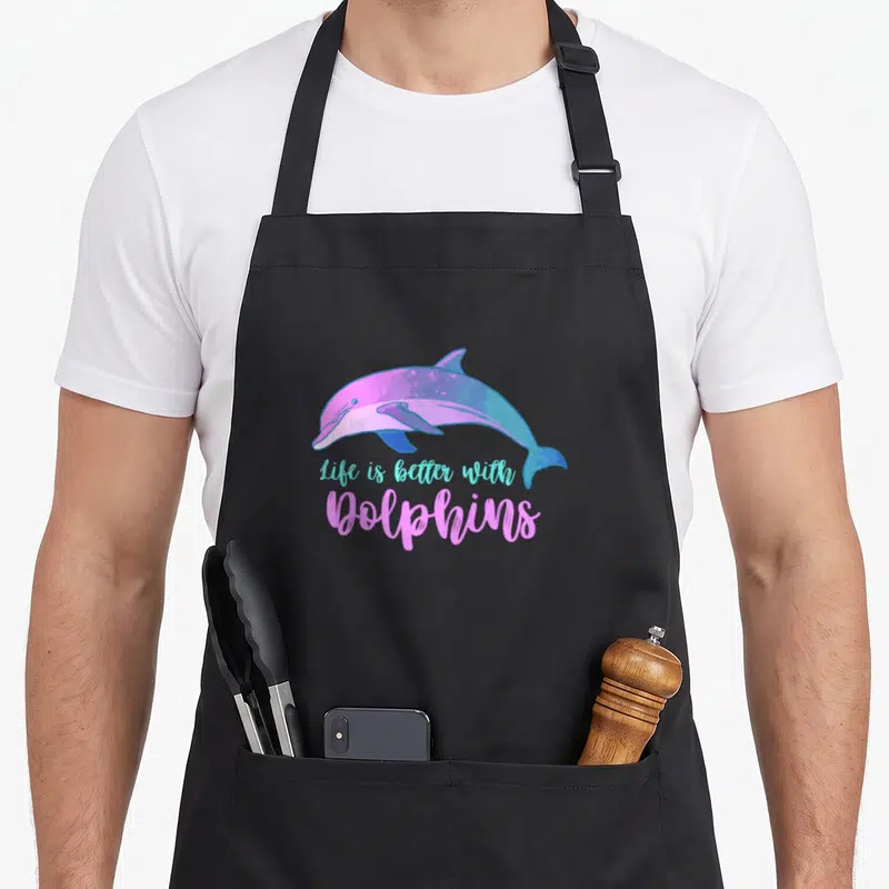 Tablier de cuisine noir avec un dauphin rose et bleu et le texte Life is better with Dolphins en typographie manuscrite.