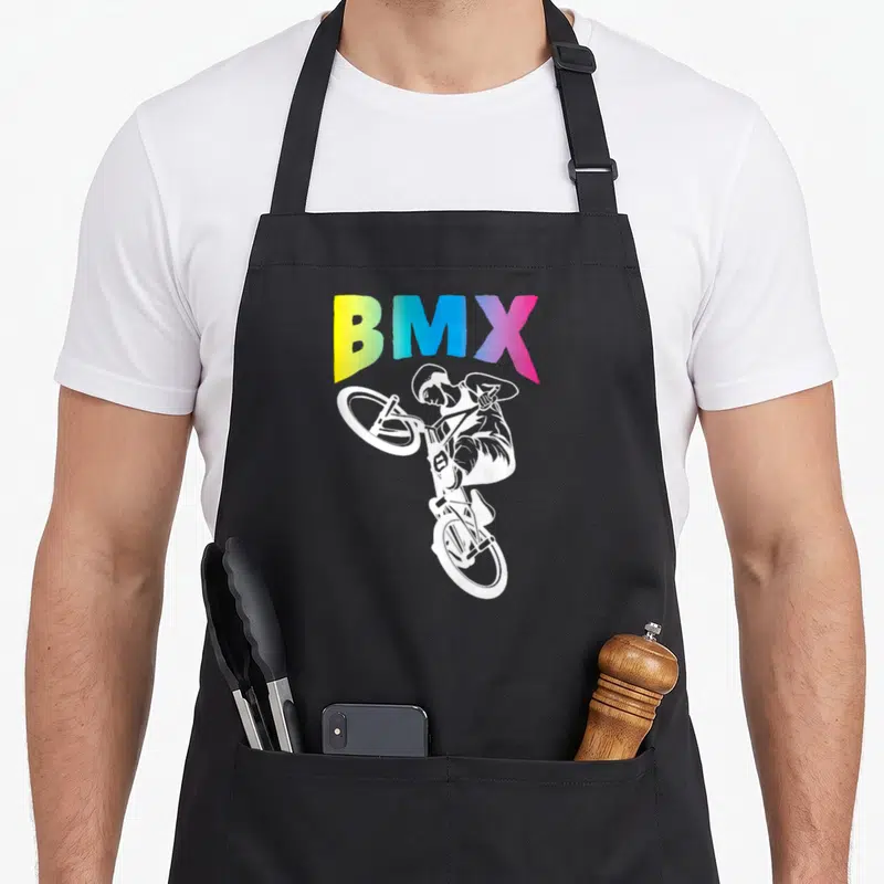 Tablier de cuisine noir avec illustration blanche d'un cycliste BMX effectuant un saut acrobatique sous un texte coloré.