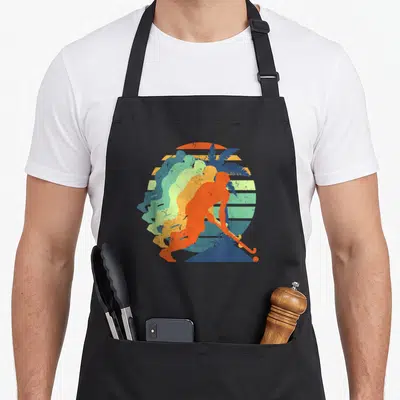 Tablier de cuisine noir avec une illustration rétro d'un joueur de hockey sur gazon orange devant un soleil rayé et des feuilles de palmier bleues.