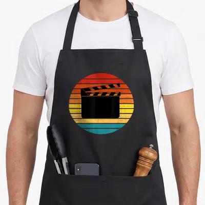 Tablier de cuisine noir avec un motif circulaire représentant un clap de film sur un fond desoleil aux couleurs chaudes et délavées.