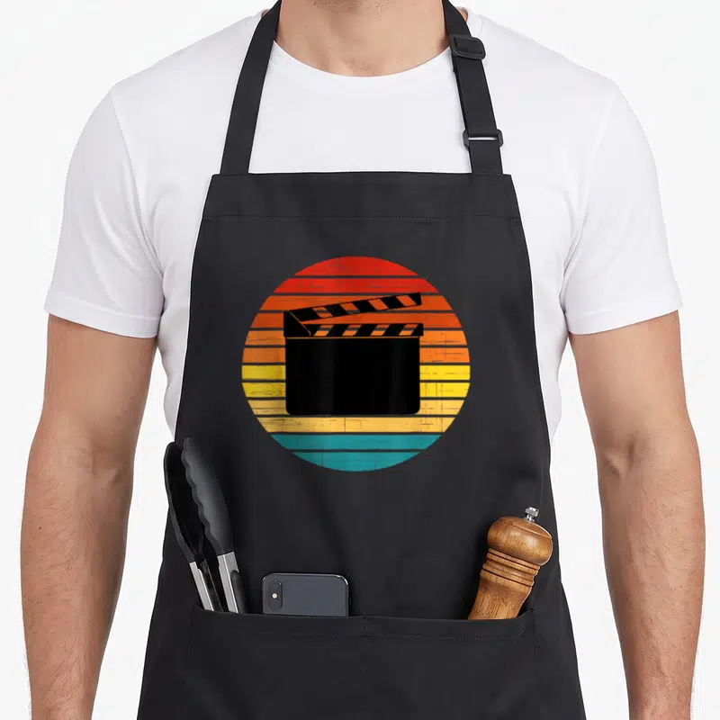 Tablier de cuisine noir avec un motif circulaire représentant un clap de film sur un fond desoleil aux couleurs chaudes et délavées.