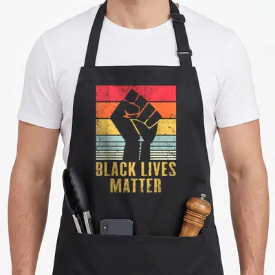 Tablier de cuisine noir avec illustration poing levé vintage et texte Black Lives Matter jaune sur bandes colorées.