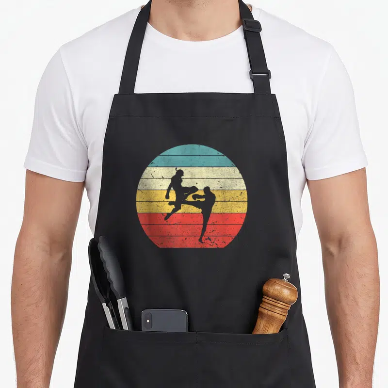 Tablier de cuisine noir avec illustration circulaire de deux silhouettes s'affrontant au Muay Thai sur fond de sunset coloré