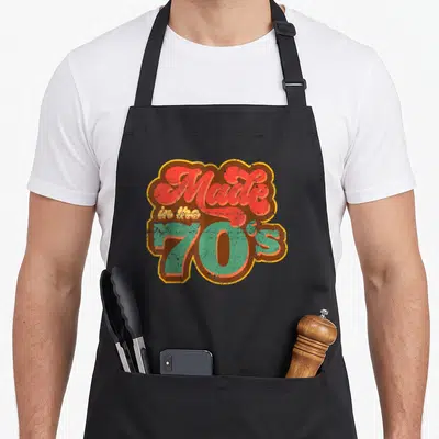 Tablier de cuisine noir avec inscription « Made in the 70s » en lettres colorées et effet vieilli.