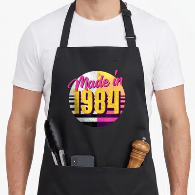 Tablier de cuisine noir avec un logo circulaire rose jaune et violet portant la mention « Made in 1984 ».