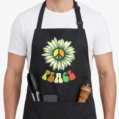 Tablier de cuisine noir avec une daisie centrale intègre un symbole de paix coloré et le texte PEACE en bas.