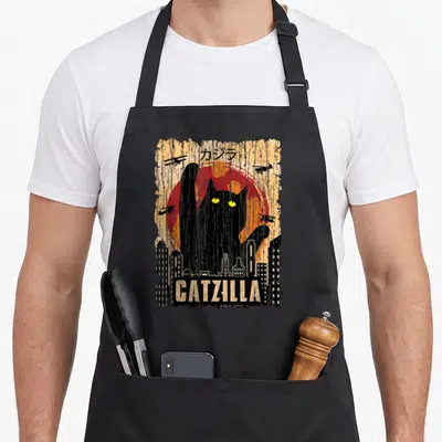 Tablier de cuisine noir avec illustration d'un chat noir géant aux yeux jaunes sur fond de soleil rouge et silhouettes de bâtiments.