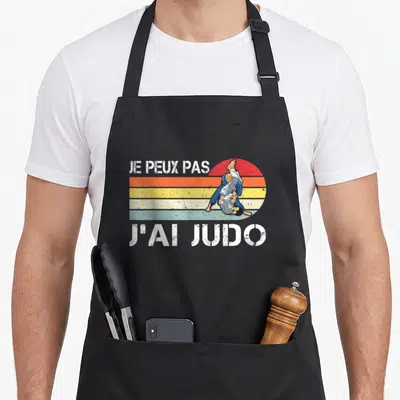 Tablier de cuisine noir avec illustration retro d'un judoka et texte blanc Je peux pas j'ai judo sur fond de soleil rayé.