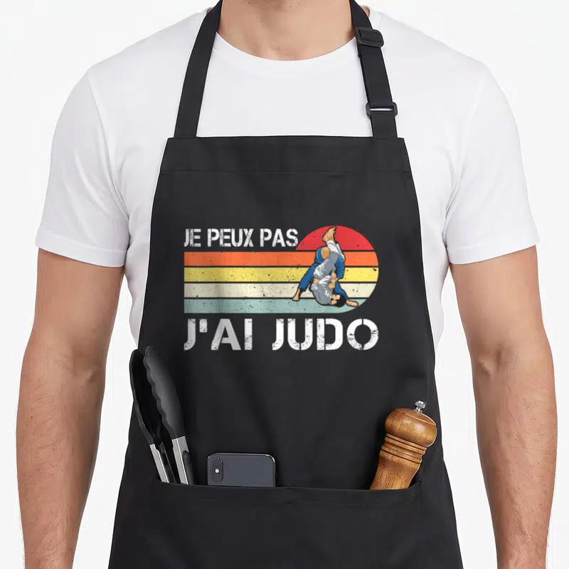 Tablier de cuisine noir avec illustration retro d'un judoka et texte blanc Je peux pas j'ai judo sur fond de soleil rayé.