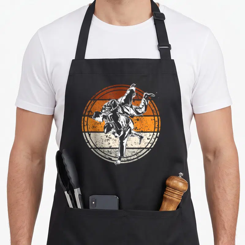 Tablier de cuisine noir avec illustration circulaire orange et blanche d'un judoiste effectuant une projection.