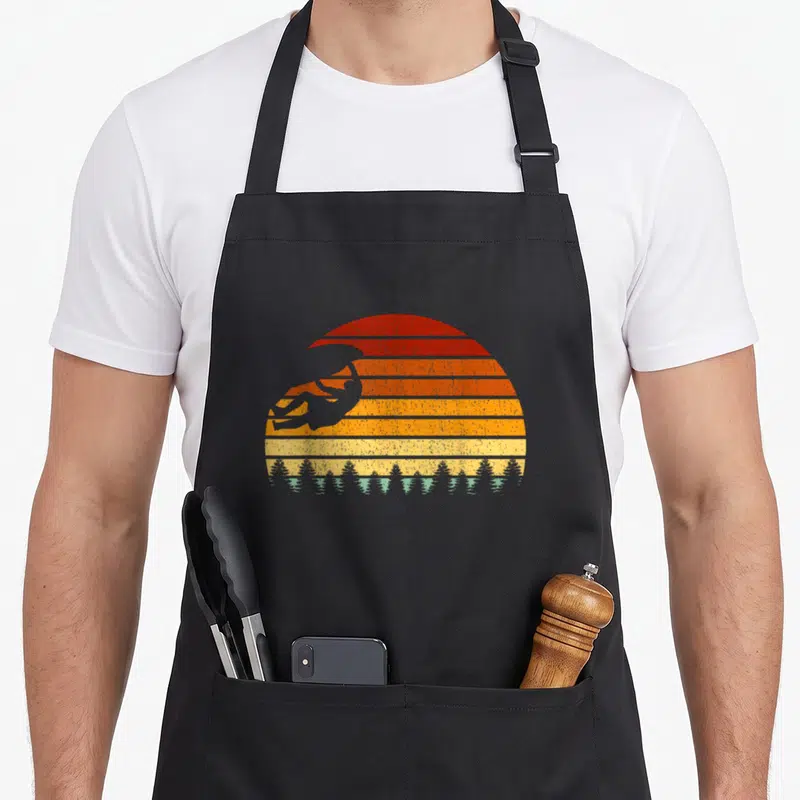 Tablier de cuisine noir avec silhouette grimpeur et soleil rayé orange retro au-dessus d'une foret d'epices.