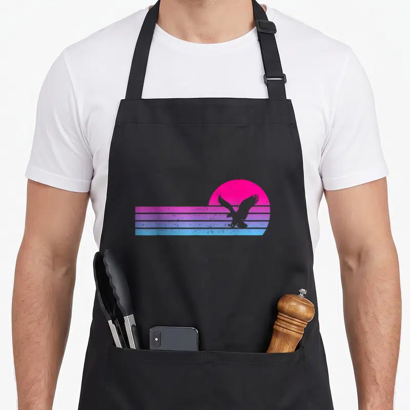 Tablier de cuisine noir avec aigle en silhouette sur fond de soleil rose et bandes horizontales bleues et violettes.