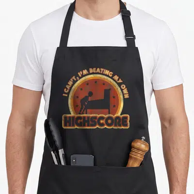 Tablier de cuisine noir avec illustration circulaire orange représentant un joueur de flipper et le texte highscore en bas.