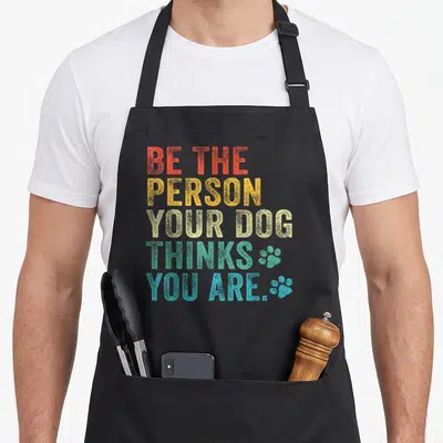 Tablier de cuisine noir avec inscription colorée Be the person your dog thinks you are et deux traces de pattes bleues en bas à droite.