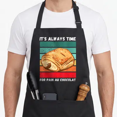 Tablier de cuisine noir avec illustration d'un pain au chocolat et texte retro sur bandes colorées.