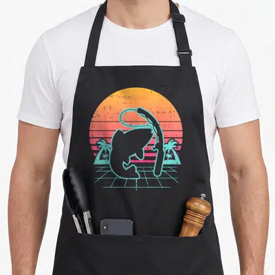 Tablier de cuisine noir avec illustration d'un poisson et d'une canne à pêche sur fond de soleil orange et palmiers turquoise.